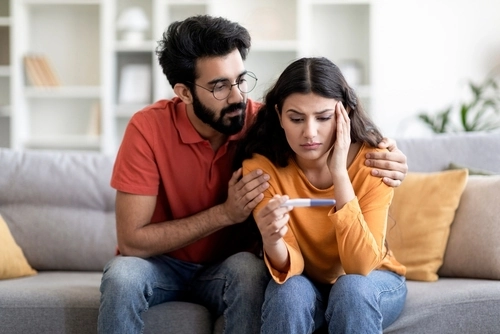 Infertility Problems. Portrait Of Upset Young Indian Couple Looking At Negative Pregnancy Test While Sitting Together On Couch At Home, Caring Eastern Husband Comforting Depressed Wife, Free Space